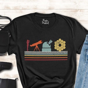 May include: Black t-shirt with a retro-style graphic of a person looking through a telescope at a star. The graphic also includes a honeycomb design and colorful stripes. The t-shirt is laid out on a wooden surface with a pair of black boots, a black purse, and blue jeans.