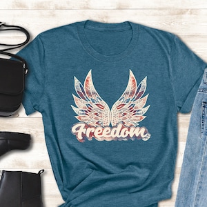 Puede incluir: Una camiseta azul turquesa con un gr&aacute;fico de alas rojas, blancas y azules con la palabra "Freedom" en una fuente vintage. La camiseta se muestra con un par de botines negros de tobillo, un bolso bandolera negro y unos vaqueros azules claros.