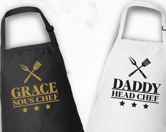 May include: Two black and white aprons with a pocket on the front. The black apron has gold text that says "Grace Sous Chef" with three gold stars. The white apron has black text that says "Daddy Head Chef" with three black stars.
