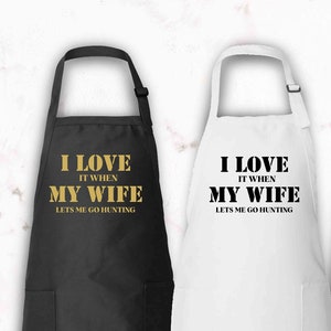 May include: Three kitchen aprons with a pocket on the bottom. The aprons are black, white, and forest green. The text on the aprons reads "I love it when my wife lets me go hunting".