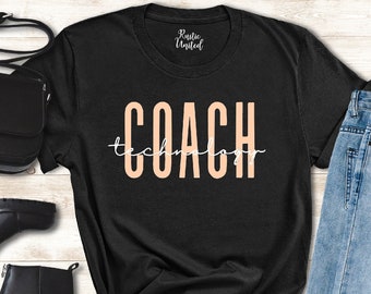 May include: Black t-shirt with the text "COACH" in peach colored lettering with the word "technology" written underneath in a smaller font. The shirt is laid out on a wooden surface with a pair of black boots, a black purse, and blue jeans.