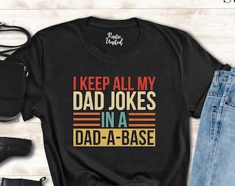 May include: Black t-shirt with the text "I KEEP ALL MY DAD JOKES IN A DAD-A-BASE" in red, orange, and yellow. The shirt is styled with black boots, a black purse, and blue jeans. Sunglasses are also included.
