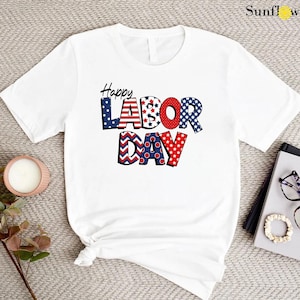 May include: White t-shirt with a red, white, and blue graphic that says "Happy Labor Day". The graphic is made up of stars, stripes, and chevron patterns.