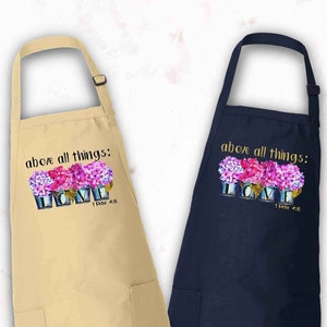 May include: Two aprons, one natural and one navy blue, are displayed. Each apron has a pocket and an adjustable neck strap. The aprons feature the text "above all things: LOVE" with a floral design. The SunflowerTeesCo logo is visible.