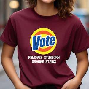 Pu&ograve; includere: Una maglietta color granata con un cerchio bianco e la parola "Vote" in lettere blu. Il cerchio ha un anello giallo e arancione intorno. Sotto il cerchio c'&egrave; il testo "Removes Stubborn Orange Stains".