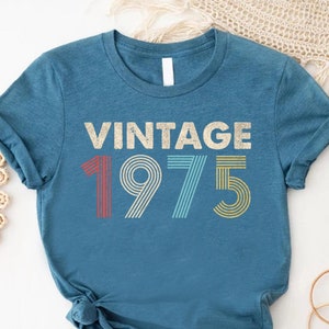 May include: A teal blue t-shirt with the text "Vintage 1975" in a retro font with colorful stripes.