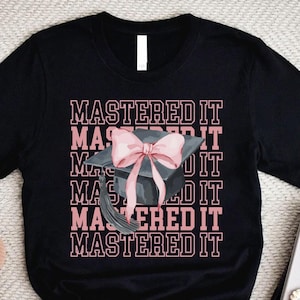 May include: Black t-shirt with the text "MASTERED IT" in pink, repeated vertically. A graduation cap with a pink bow is centered on the design. The shirt is displayed on a neutral surface with a candle, glasses, and a book.