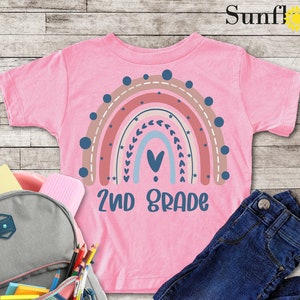 May include: A pink t-shirt with a rainbow design and the text "2nd Grade". The rainbow is made up of different shades of pink, orange, and blue. There are small hearts and dots around the rainbow. The shirt is laid out on a wooden surface with a backpack, pencils, and jeans.
