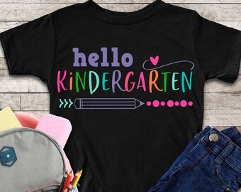May include: Black t-shirt with the text "hello kindergarten" in rainbow colours, a pink heart, a pencil, and dots. The shirt is laid out on a wooden surface with a backpack, jeans, and school supplies.