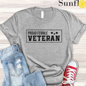 May include: Light grey heather t-shirt with the text "Proud Female Veteran" in a black rectangle with three stars above the text. The t-shirt is laying on a wood surface with a pair of blue jeans and red sneakers.