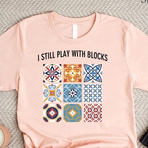 May include: A light pink t-shirt with the text "I still play with blocks" and a graphic of nine colorful patterned tiles.