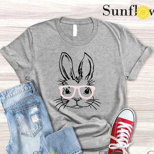 May include: A light grey heather t-shirt with a black and white illustration of a bunny wearing pink glasses. The t-shirt is laid flat on a wooden surface with a pair of blue jeans and red sneakers.