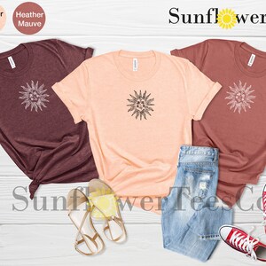 May include: Three t-shirts with a sun graphic design in maroon, peach, and rust colors. The shirts are laid out on a wooden surface with a pair of sandals and jeans. The text "Sunflower TeesCo" is visible at the top of the image.