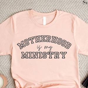May include: A light pink t-shirt with the text "MOTHERHOOD is my MINISTRY" in a black collegiate font.