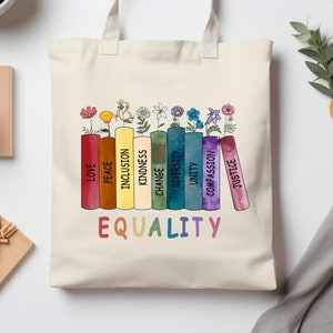 May include: A white canvas tote bag with a rainbow-colored design of stacked books. Each book has a word printed on the spine, including "Love", "Peace", "Inclusion", "Kindness", "Change", "Diversity", "Unity", "Compassion", and "Justice". The words are arranged in a rainbow pattern. The words "EQUALITY" are printed in rainbow colors below the books.