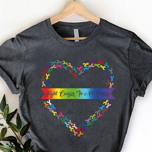 May include: Dark gray t-shirt with a heart-shaped design made of colorful ribbons. A rainbow banner across the heart reads "Fight Cancer In All Colors". The shirt is displayed on a wooden hanger against a white background.