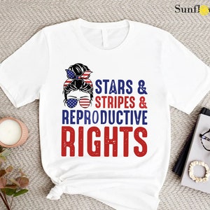 May include: White t-shirt with a red, white, and blue graphic of a woman with a bun in her hair wearing sunglasses and a headband. The text "Stars & Stripes & Reproductive Rights" is printed in red and blue.
