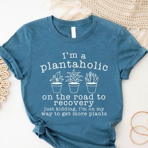 Op de afbeelding: Een tealblauw t-shirt met een witte afbeelding waarop staat "Ik ben een plantaholic op weg naar herstel, grapje, ik ben op weg om meer planten te halen".