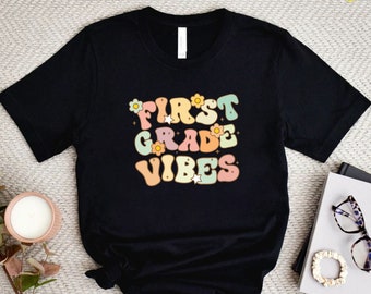 May include: Black t-shirt with the text "First Grade Vibes" in colorful retro letters with flowers and stars.