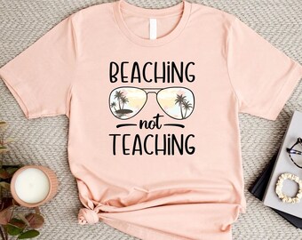 May include: A pink t-shirt with a graphic of sunglasses with palm trees reflected in the lenses. The text "Beaching - not teaching" is printed on the shirt.