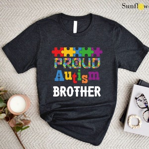 May include: Dark grey t-shirt with a white graphic design. The graphic features a row of colorful puzzle pieces that spell out "Proud Autism Brother".