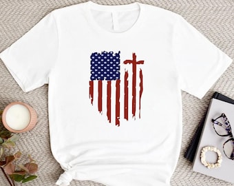 Puede incluir: Camiseta blanca con un diseño de bandera estadounidense desgastada y una cruz roja en el centro.