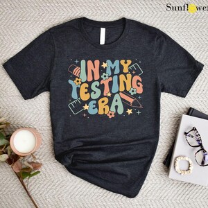 May include: A dark gray t-shirt with the text "In My Testing Era" in a retro font with colorful accents. The text is surrounded by stars, pencils, and rulers.