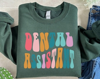 May include: A dark green sweatshirt with the words "Dental Assistant" in colorful, wavy letters. The letters are in shades of pink, orange, yellow, green, and blue.