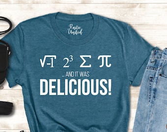 May include: A teal blue t-shirt with white text that reads "√-1 2^3 Σ π AND IT WAS DELICIOUS!" The shirt is laid out on a wooden surface with a pair of black boots, a black purse, and blue jeans.
