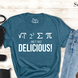 May include: A teal blue t-shirt with white text that reads "√-1 2^3 Σ π AND IT WAS DELICIOUS!" The shirt is laid out on a wooden surface with a pair of black boots, a black purse, and blue jeans.