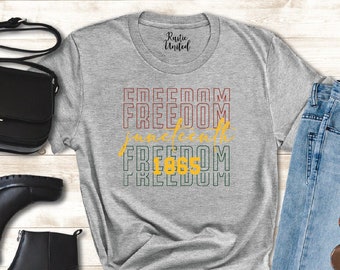 May include: Gray t-shirt with the text "FREEDOM" repeated four times in a retro style with yellow and red accents. The text "American" is in between the second and third "FREEDOM" text. The shirt is displayed on a wooden background with a pair of black boots, a black purse, and blue jeans with sunglasses.