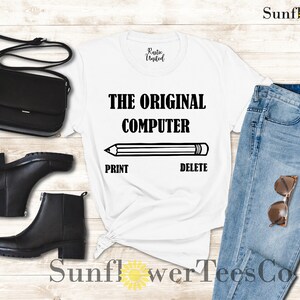 K&ouml;nnte beinhalten: Wei&szlig;es T-Shirt mit schwarzem Text, der "THE ORIGINAL COMPUTER" lautet, mit einer Bleistiftgrafik und den W&ouml;rtern "PRINT" und "DELETE" darunter. Das T-Shirt wird auf einem Holzgrund mit einer schwarzen Handtasche, schwarzen Stiefeln, einer blauen Jeans und einer Sonnenbrille gezeigt.