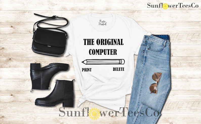 K&ouml;nnte beinhalten: Wei&szlig;es T-Shirt mit schwarzem Text, der "THE ORIGINAL COMPUTER" lautet, mit einer Bleistiftgrafik und den W&ouml;rtern "PRINT" und "DELETE" darunter. Das T-Shirt wird auf einem Holzgrund mit einer schwarzen Handtasche, schwarzen Stiefeln, einer blauen Jeans und einer Sonnenbrille gezeigt.