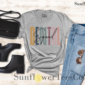 May include: A gray t-shirt with the text "Dental Squad" in colorful stripes. The shirt is styled with black boots, a black purse, and blue jeans.