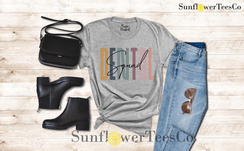 May include: A gray t-shirt with the text "Dental Squad" in colorful stripes. The shirt is styled with black boots, a black purse, and blue jeans.