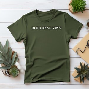 K&ouml;nnte beinhalten: Ein gr&uuml;nes T-Shirt mit dem Aufdruck "IS HE DEAD YET?" in wei&szlig; auf der Vorderseite.