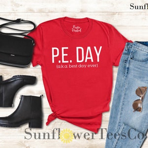 Op de afbeelding: Rood T-shirt met witte tekst die "P.E. DAY (a.k.a. best day ever)" luidt. Het shirt is gestyled met een blauwe jeans, zwarte laarzen en een zwarte handtas.