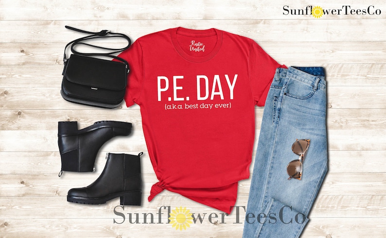 Op de afbeelding: Rood T-shirt met witte tekst die "P.E. DAY (a.k.a. best day ever)" luidt. Het shirt is gestyled met een blauwe jeans, zwarte laarzen en een zwarte handtas.