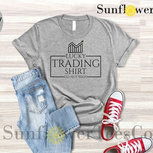 May include: A light grey heather t-shirt with black text that reads "Lucky Trading Shirt - Do Not Wash". The shirt is laid flat on a wooden surface with a pair of blue jeans and red sneakers.