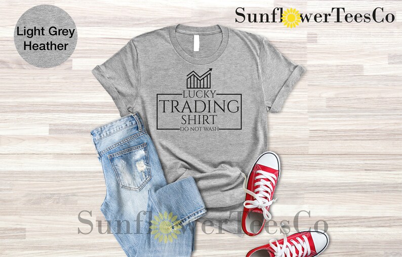 May include: A light grey heather t-shirt with black text that reads "Lucky Trading Shirt - Do Not Wash". The shirt is laid flat on a wooden surface with a pair of blue jeans and red sneakers.
