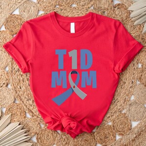 Pu&ograve; includere: Maglietta rossa con il testo "T1D MOM" in lettere blu e grigie. Un nastro blu e grigio &egrave; dietro il testo.