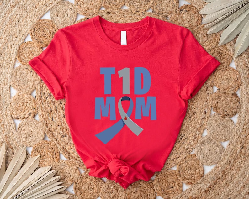Pu&ograve; includere: Maglietta rossa con il testo "T1D MOM" in lettere blu e grigie. Un nastro blu e grigio &egrave; dietro il testo.