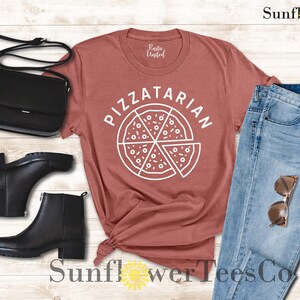 May include: A rust-colored t-shirt with the text "PIZZATARIAN" and a white outline of a pizza slice. The shirt is laid out on a wooden surface with a black purse, black boots, and blue jeans.