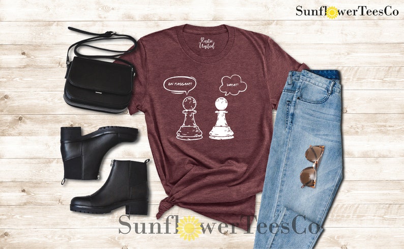May include: A maroon t-shirt with a white graphic of two chess pieces. The white piece has a thought bubble above it that says "What?!" and the black piece has a thought bubble above it that says "En Passant!"
