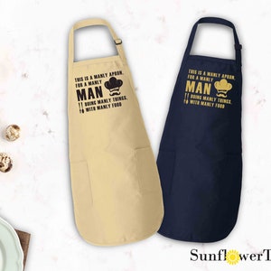 May include: Two aprons with a pocket on the bottom. The apron on the left is beige with black text that reads "This is a manly apron for a manly man doing manly things with manly food". The apron on the right is navy blue with gold text that reads "This is a manly apron for a manly man doing manly things with manly food".