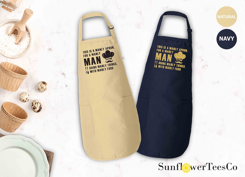 May include: Two aprons with a pocket on the bottom. The apron on the left is beige with black text that reads "This is a manly apron for a manly man doing manly things with manly food". The apron on the right is navy blue with gold text that reads "This is a manly apron for a manly man doing manly things with manly food".