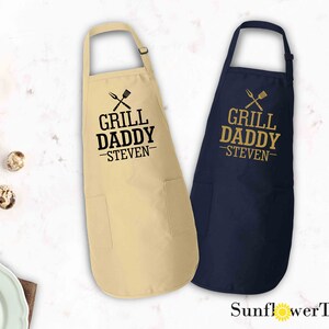 May include: Two personalized aprons with the text "Grill Daddy - Steven" in gold lettering. The aprons are available in natural and navy blue.