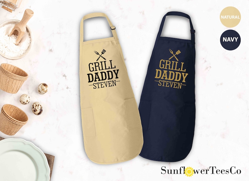 May include: Two personalized aprons with the text "Grill Daddy - Steven" in gold lettering. The aprons are available in natural and navy blue.