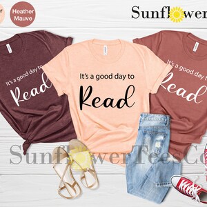 Op de afbeelding: Drie t-shirts in heather maroon, perzik en mauve, elk met de tekst "It's a good day to Read". Het perzikkleurige shirt wordt gecombineerd met een spijkerbroek en sandalen, terwijl het mauve shirt is gestyled met een spijkerbroek en rode sneakers.