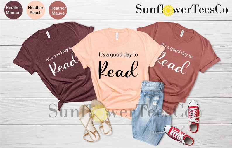 Op de afbeelding: Drie t-shirts in heather maroon, perzik en mauve, elk met de tekst "It's a good day to Read". Het perzikkleurige shirt wordt gecombineerd met een spijkerbroek en sandalen, terwijl het mauve shirt is gestyled met een spijkerbroek en rode sneakers.
